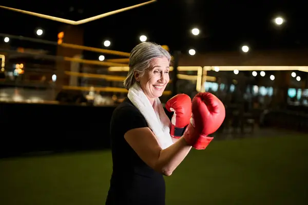 Mujer atractiva madura alegre con pelo gris y guantes de boxeo haciendo ejercicio activamente mientras que en el gimnasio - foto de stock