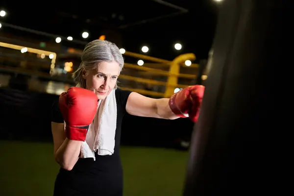 Attraktive sportliche Seniorin mit grauen Haaren und Handtuch in Boxhandschuhen schlägt Boxsack in Turnhalle — Stockfoto