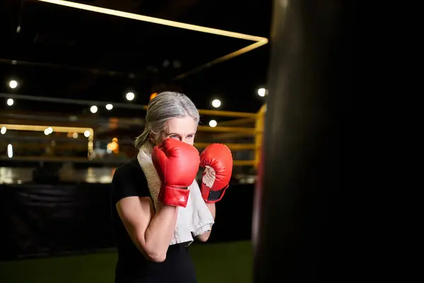Mujer madura de buen aspecto con pelo gris y toalla en guantes de boxeo golpeando saco de boxeo en el gimnasio - foto de stock