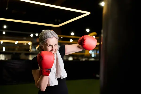 Athletic beautiful senior woman with gray hair in boxing gloves beating punching bag in gym — Stock Photo
