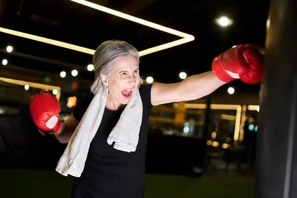 Atractiva mujer madura deportiva con pelo gris y toalla en guantes de boxeo golpeando saco de boxeo en el gimnasio - foto de stock