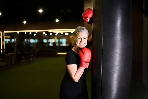 Jolly senior woman in sportswear posing with boxing gloves near punching bag and smiling at camera — Stock Photo