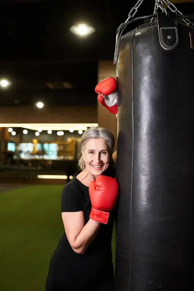 Sportif senior joyeux sportif posant avec des gants de boxe près du sac de boxe et souriant à la caméra — Photo de stock