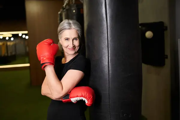 Joyous mature woman in sportswear posing with boxing gloves near punching bag and smiling at camera — Stock Photo