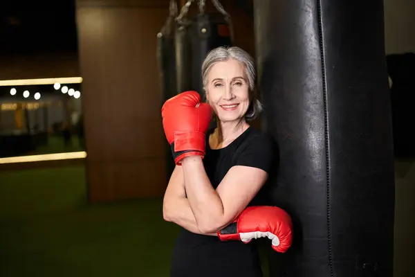 Mature athletic jolly sportswoman posing with boxing gloves near punching bag and smiling at camera — Stock Photo
