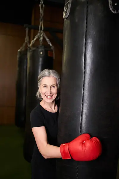 Allegra donna matura in abbigliamento sportivo con guanti da boxe vicino al sacco da boxe e sorridente alla macchina fotografica — Foto stock