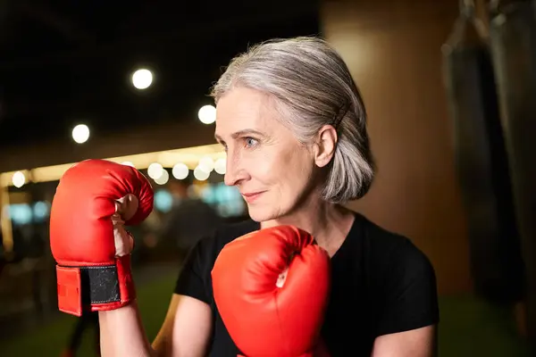 Sportif belle femme âgée avec des cheveux gris posant avec des gants de boxe tout en s'entraînant dans la salle de gym — Photo de stock