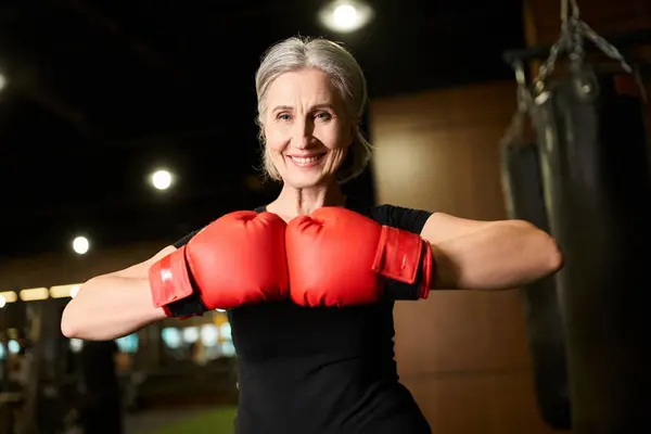 Joyful pretty mature sportswoman with gray hair in boxing gloves smiling at camera while in gym — Stock Photo