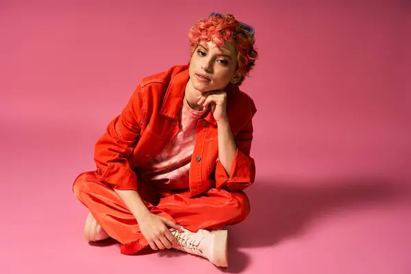 Uma mulher com cabelo vermelho em trajes vibrantes senta-se elegantemente em um fundo rosa. — Fotografia de Stock