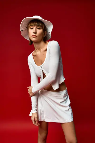 Elegante Frau in weißer Kleidung und Hut posiert anmutig vor leuchtend rotem Hintergrund. — Stockfoto