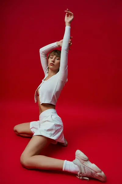 Junge Frau in eleganter weißer Sportbekleidung sitzt mit erhobenen Armen in freudiger Pose auf dem Boden. — Stockfoto