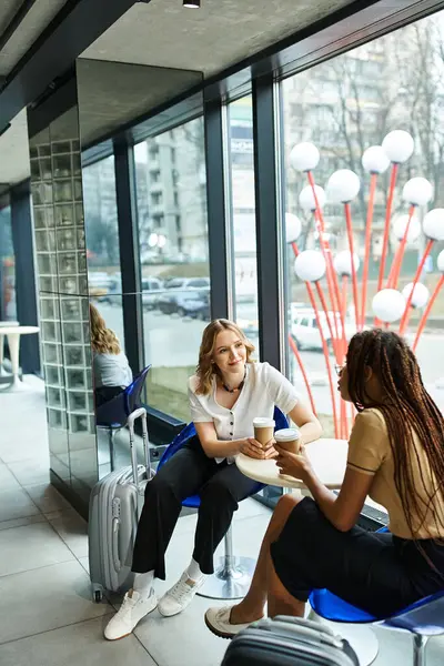 Two multicultural colleagues, diverse businesswomen, sitting at a table in a hotel, engaged in deep conversation in front of a window. — Stock Photo