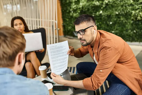 Un homme, qui fait partie d'une équipe d'affaires moderne, s'assoit à une table tenant un bout de papier tout en étant entouré de collègues dans un espace de coworking. — Photo de stock