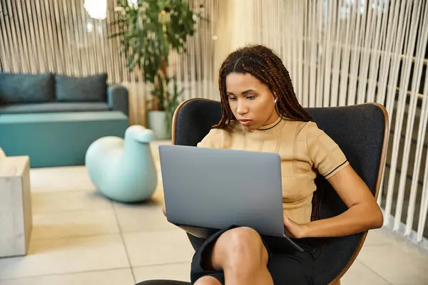 Una donna si siede su una sedia, focalizzata sul suo schermo portatile, lavorando in un ambiente di business moderno. — Foto stock