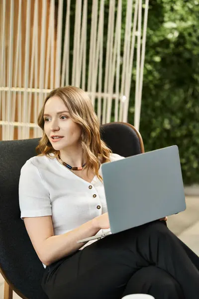 Una donna si siede su una sedia, intensamente concentrata sul suo computer portatile, immersa nella moderna vita d'affari con i colleghi in uno spazio di coworking. — Foto stock