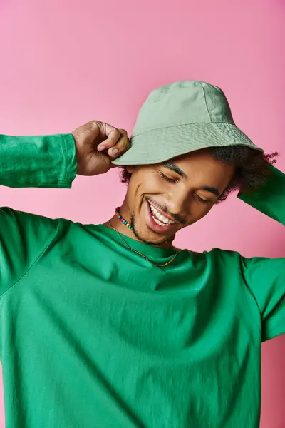 A cheerful young African American man with curly hair wearing a green shirt and green hat on a pink background. — Stock Photo