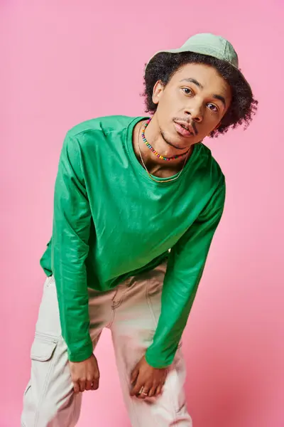 Ein fröhlicher, junger und lockiger afroamerikanischer Mann in grünem Hemd und weißer Hose vor rosa Hintergrund. — Stockfoto