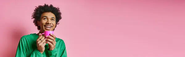 Hombre afroamericano rizado con camisa verde sostiene alegremente una copa rosa brillante sobre un fondo rosa. - foto de stock