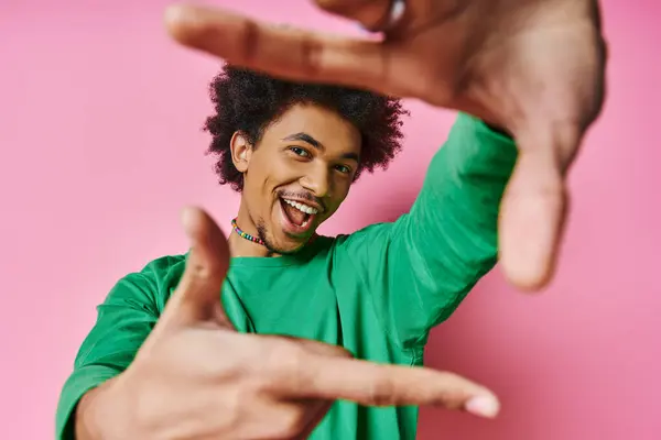 Un joven y alegre afroamericano en ropa casual sobre un fondo rosado hace un gesto con sus manos, mostrando varias emociones. - foto de stock