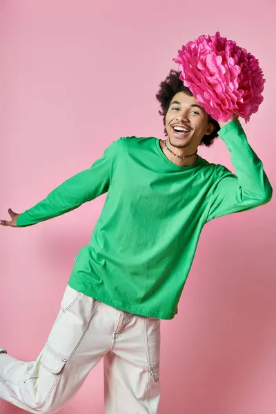Веселый молодой афроамериканец с вьющимися волосами, держащий розовый цветок в зеленой рубашке на розовом фоне. — стоковое фото