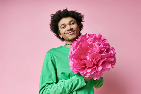 Un homme afro-américain joyeux et bouclé en tenue décontractée tient une grande fleur rose sur un fond rose. — Photo de stock