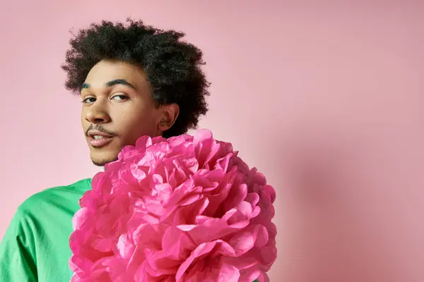 Ein fröhlicher junger afroamerikanischer Mann in lässiger Kleidung hält sich eine große rosafarbene Blume vor das Gesicht, die Emotionen und Eleganz zur Schau stellt. — Stockfoto
