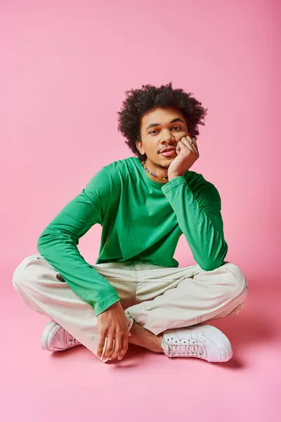 Young African American man with curly hair wearing a green shirt and white pants on a pink background. — Stock Photo