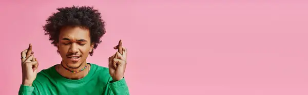 Hopeful young African American man in casual wear holds up two fingers in front of his face against a pink background. — Stock Photo
