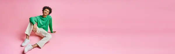 A cheerful African American man in a green shirt and white pants sits on a pink background, exuding emotion and charm. — Stock Photo