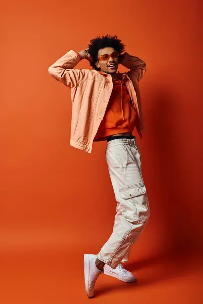Ein modischer junger afroamerikanischer Mann mit lockigem Haar und Sonnenbrille posiert selbstbewusst vor leuchtend orangefarbener Kulisse. — Stockfoto