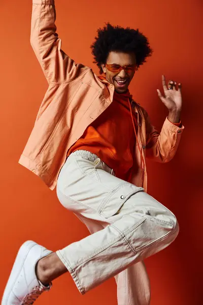 Curly African American man in trendy outfit and sunglasses, jumping energetically in the air against orange background. — Stock Photo