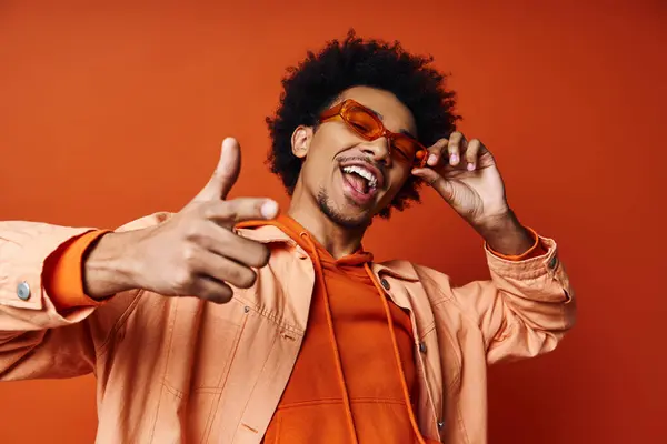 Стильний молодий афроамериканський чоловік в помаранчевій сорочці та сонцезахисних окулярах робить дурне обличчя на помаранчевому фоні. — стокове фото