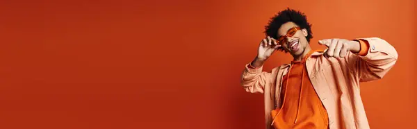 A stylish young African American man in an orange shirt and sunglasses holding his hand to his face on a vibrant orange background. — Stock Photo