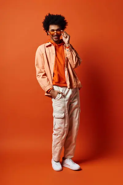 A trendy, emotional young African American man stands confidently against a bright orange background in fashionable attire and sunglasses. — Stock Photo