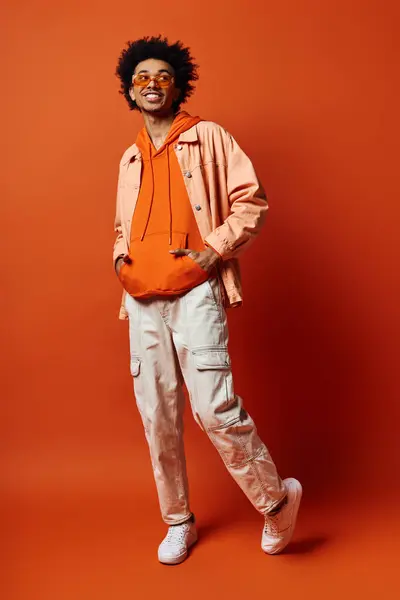 Jeune homme afro-américain en tenue tendance et lunettes de soleil, exprimant des émotions sur fond orange. — Photo de stock