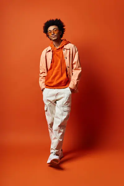 Jeune homme afro-américain élégant en tenue tendance et lunettes de soleil respirant l'émotion devant une toile de fond orange frappante. — Photo de stock