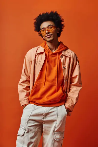 Stilvoller junger Afroamerikaner in orangefarbenem Kapuzenpulli und weißer Hose, der Zuversicht und Emotion ausstrahlt. — Stockfoto