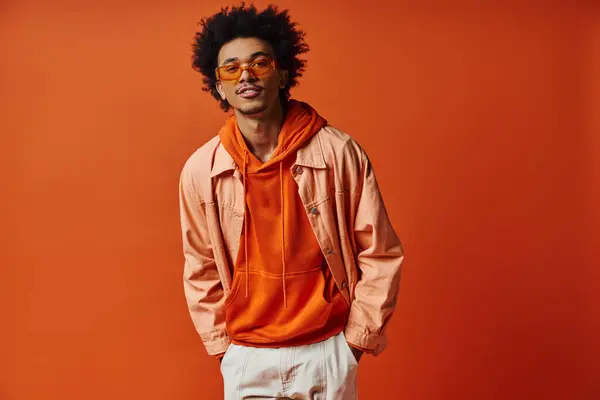 Um homem afro-americano encaracolado exalando estilo e emoção na camisa laranja na moda, shorts brancos e óculos de sol em um fundo laranja. — Fotografia de Stock