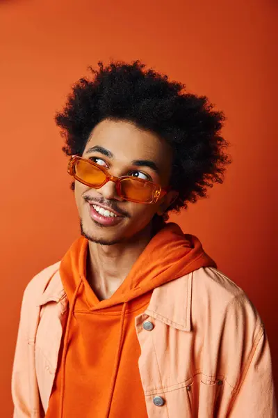 A fashionable African American man wearing a trendy jacket and sunglasses, exudes confidence against an orange background. — Stock Photo