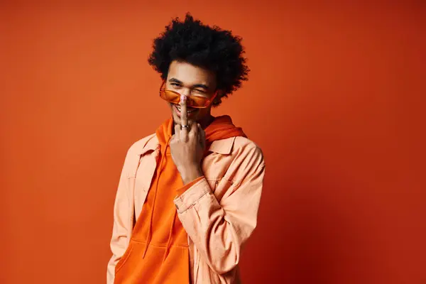 Elegante jovem afro-americano, cabelo encaracolado, camisa laranja, mãos no rosto, expressando emoção, fundo na moda. — Fotografia de Stock