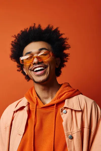 Stilvoller junger afroamerikanischer Mann in orangefarbenem Kapuzenpulli und Sonnenbrille mit lockigem Afro-Haar auf leuchtend orangefarbenem Hintergrund, der Emotionen ausdrückt. — Stockfoto