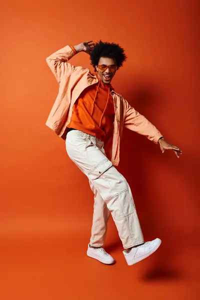 Ein angesagter afroamerikanischer Mann mit lockigem Haar und Sonnenbrille posiert in einem orangefarbenen Hemd und einer weißen Hose vor orangefarbenem Hintergrund. — Stockfoto