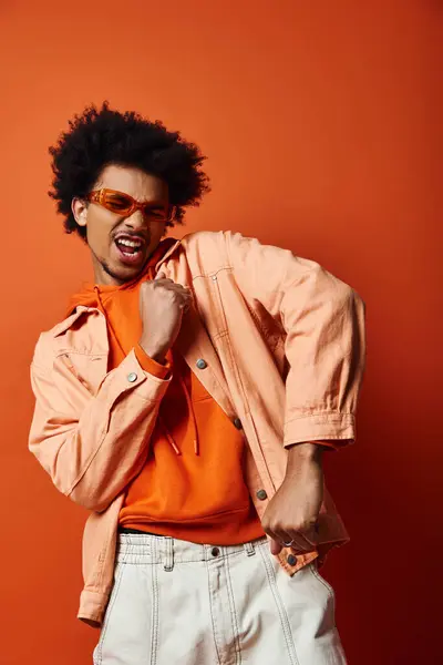 Stylish African American man in orange shirt and jacket, wearing sunglasses, on orange background. Trendy and expressive. — Stock Photo
