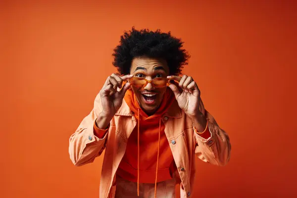 A stylish, young African American man in an orange jacket holds a pair of glasses against an orange background. — Stock Photo