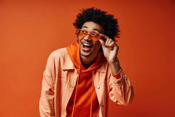 A young African American man with curly hair in trendy attire and sunglasses, showcasing emotions against an orange background. — Stock Photo