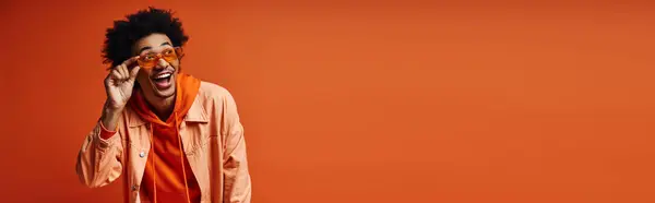Ein stilvoller junger afroamerikanischer Mann mit lockigem Haar und modischer Kleidung verhüllt seine Augen mit einem orangefarbenen Schal vor orangefarbenem Hintergrund.. — Stockfoto