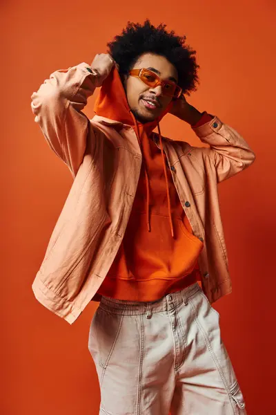 A stylish young African American man with curly hair wearing an orange shirt and white shorts, exuding confidence on an orange background. — Stock Photo