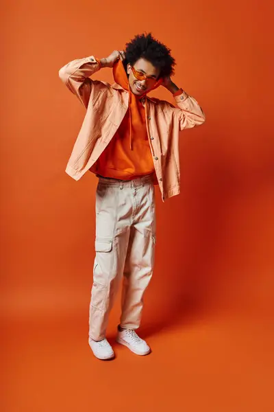 Hombre afroamericano rizado con camisa naranja de moda y pantalones caqui, estilo exuberante y carisma sobre un fondo naranja audaz. - foto de stock