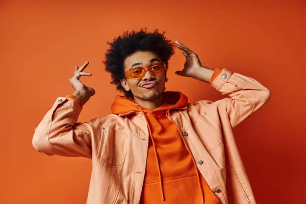 Stylish African American man in orange jacket and glasses against a bold orange background. — Stock Photo