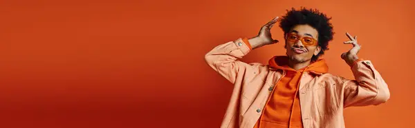 Un giovane afroamericano elegante con i capelli ricci che indossa una camicia arancione, pantaloni e occhiali da sole, in posa con un'espressione emotiva. — Foto stock